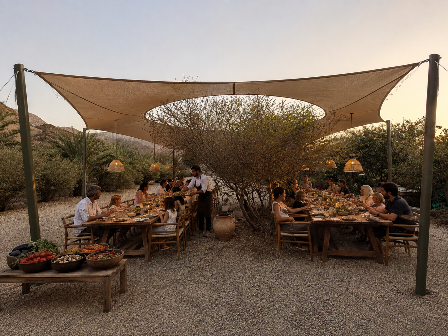 Guests dining at a long chef's table under shade sails at the farm at sunset